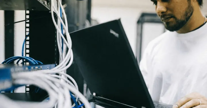 Technician working on a laptop beside a server rack full of cables